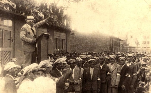 Rumkowski stands on a podium, speaking to a large crowd of mostly men. He has his left arm raised in a gesture similar to a Nazi salute.